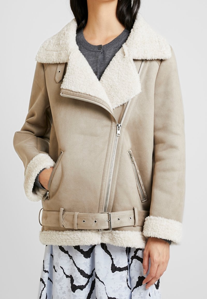 Beige shearling-lined jacket with a zip closure, wide lapels, and adjustable belt. Features side pockets and contrasting textured cuffs.
