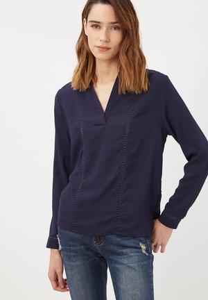 Blusa de manga larga azul marino con escote en V, que presenta costuras verticales y detalles en el escote y puños. Llevada con jeans de mezclilla.
