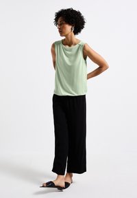 Blusa senza maniche verde chiaro con una vestibilità rilassata, abbinata a pantaloni neri a gamba larga e sandali neri slip-on su uno sfondo neutro.