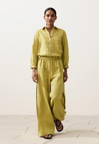 Femeie care merge purtând o cămașă largă verde deschis cu nasturi, asortată cu pantaloni evazați și papuci negri, pe un podea cu plăci deschise la culoare.