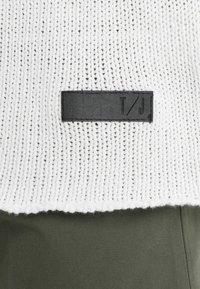 Bílý pletený svetřík s texturovaným vzorem, s černou koženou cedulkou s nápisem "T/J" přišitou na dolním okraji.