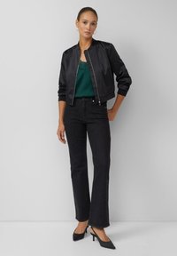 Veste bomber en satin noir, caraco en dentelle verte, jean évasé foncé, et escarpins noirs à bouts pointus. Design simple avec des textures contrastantes.