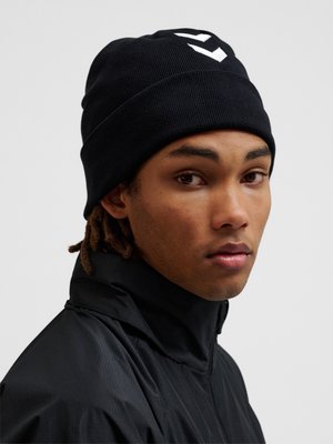 En ung person med mellemtonet hudfarve iført en sort strikket hue med hvid chevron-logo og en sort jakke, der kigger let over skulderen.