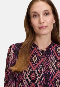 Long-sleeved blouse with a colorful geometric pattern in pink, black, and cream. Features a keyhole neckline and button accents.