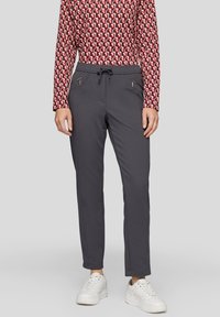 Graue Hose mit elastischem Bund, zwei Reißverschlusstaschen und schmal zulaufendem Schnitt, kombiniert mit einem gemusterten Langarmoberteil und weißen Sneakern.