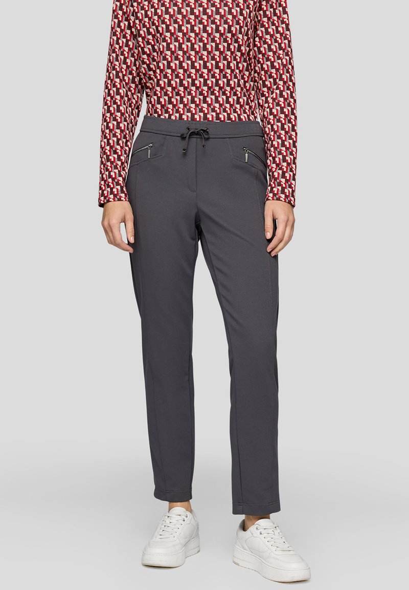 Graue Hose mit elastischem Bund, zwei Reißverschlusstaschen und schmal zulaufendem Schnitt, kombiniert mit einem gemusterten Langarmoberteil und weißen Sneakern.