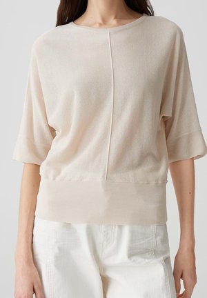 Light beige short-sleeve top with a loose fit, ribbed hem, and central seam detail, made from a soft, textured fabric. Paired with white pants.