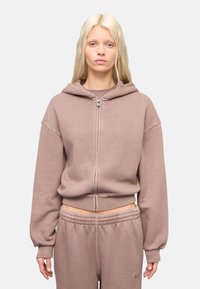 Sudadera con capucha de cremallera en marrón claro, hecha de tejido suave. Presenta mangas amplias y una cinturilla acanalada. Combinada con pantalones de chándal a juego.