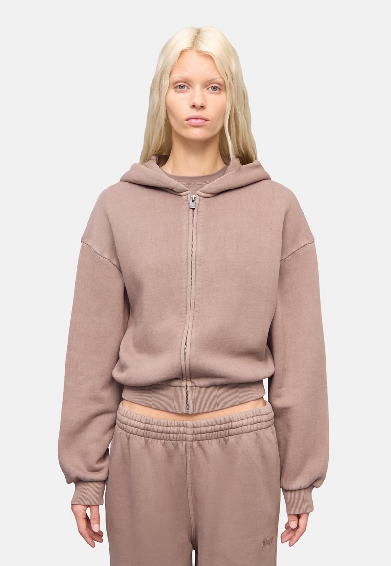Sudadera con capucha de cremallera en marrón claro, hecha de tejido suave. Presenta mangas amplias y una cinturilla acanalada. Combinada con pantalones de chándal a juego.