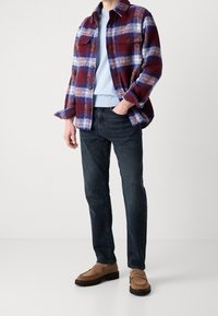Hombre vistiendo una chaqueta de cuadros en rojo, blanco y azul, camisa celeste, jeans oscuros y mocasines marrones, de pie frente a un fondo liso.