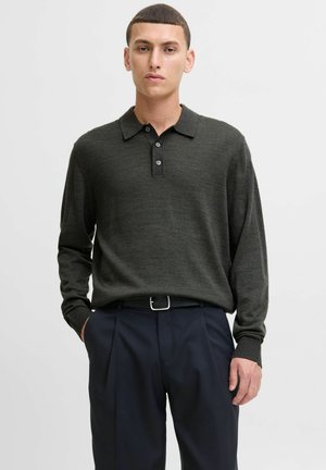 Joven con el pelo corto que lleva un suéter de polo de manga larga verde oscuro metido en unos pantalones negros de cintura alta con cinturón.