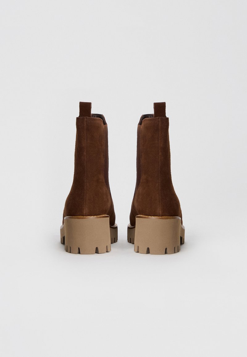 Brązowe zamszowe botki sięgające kostki z grubą beżową podeszwą, z gładkim wykończeniem i pętelką z tyłu ułatwiającą zakładanie.