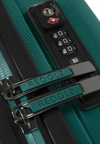 Valigia verde con texture, dotata di lucchetto TSA a combinazione nera e doppi cursori della zip incisi con "REDOLZ" in primo piano.