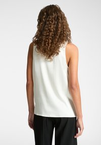 Top bianco senza maniche realizzato in tessuto morbido, con una vestibilità comoda e orlo arrotondato, abbinato a pantaloni neri. Capelli ricci in mostra.