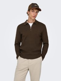 Maglione marrone a maglia con colletto e mezza cerniera frontale, abbinato a pantaloni beige. Il modello indossa un berretto da baseball marrone con la scritta "Detroit".