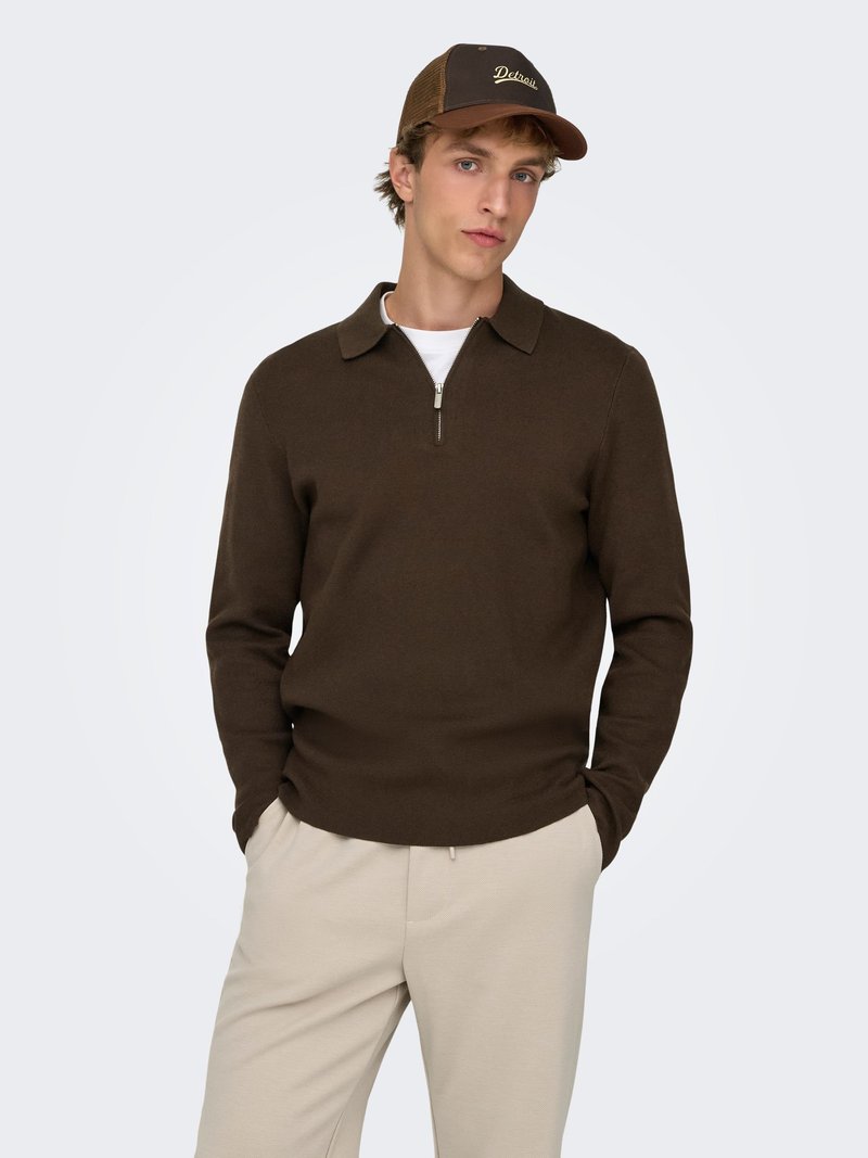 Maglione marrone a maglia con colletto e mezza cerniera frontale, abbinato a pantaloni beige. Il modello indossa un berretto da baseball marrone con la scritta "Detroit".