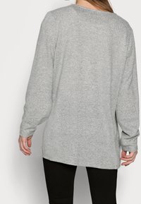 Suéter gris de manga larga con una tela texturada, un escote redondeado y un dobladillo trasero ligeramente más largo, combinado con pantalones negros.