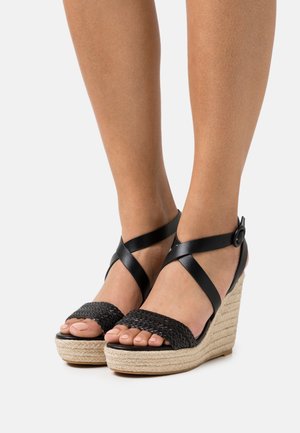 Sandales compensées noires avec une bride tissée, une boucle à la cheville et une semelle en jute tressé. Présentent des brides croisées pour un soutien et un style supplémentaires.