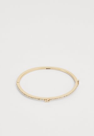 Zlati ton bangle zapestnica z gladko površino, okrašena s prozornimi kamnitimi kamenčki ob strani in z zapiranjem na sponko. Logotip viden znotraj.