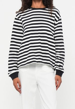 Persona vistiendo una camisa de manga larga con rayas horizontales negras y blancas, puños negros y pantalones blancos, mostrada desde el cuello hasta las caderas.