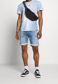 Musta kangaskotelo vyötäröllä, vaaleansininen t-paita, jossa on sitruunapainatus, käänteiset denim-shortsit ja mustat Vans-lenkkarit, joissa on valkoiset pohjat.