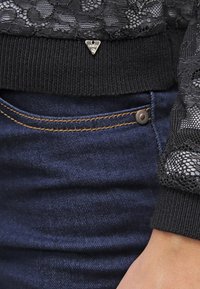 Haut en dentelle noire avec ourlet côtelé, orné d'un petit logo en métal. Associé à un jean en denim foncé, souligné par des coutures contrastées et un bouton.