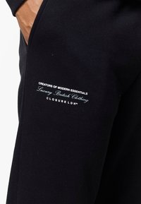 Hand in de zak van zwarte broek met witte en lichtblauwe tekst op het dijbeen met de woorden "Creators of Modern Essentials Luxury British Clothing Closure LDN™".