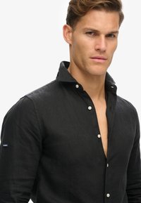 Camisa de lino negra con cuello abotonado, que presenta botones blancos y una pequeña etiqueta de marca en la manga. La tela texturizada ofrece un aspecto relajado.