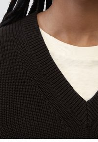 Schwarzer Strickpullover mit tiefem V-Ausschnitt, über einem cremefarbenen Shirt mit weicher Textur getragen.
