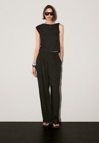 Black sleeveless top with draped neckline, paired with wide-leg black trousers. Model wears sunglasses and bracelets. Smooth fabric texture.
