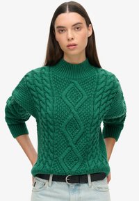 Pull en maille verte avec des motifs en relief, un col montant et des poignets côtelés. Porté avec un jean bleu clair et une ceinture noire.