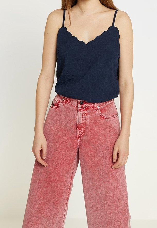 Femme portant un top sans manches bleu marine à bord festonné et un jean rose taille haute à jambes larges sur un fond uni.