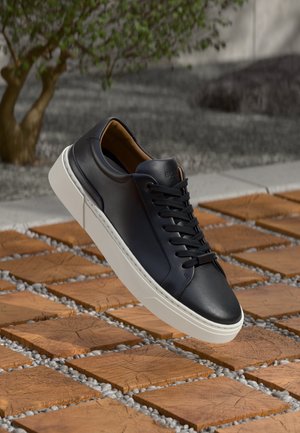 Czarne skórzane sneakersy z białą podeszwą, z okrągłymi sznurówkami, smukłym designem i brązowym wnętrzem. Wyeksponowane na wzorzystych drewnianych płytkach.