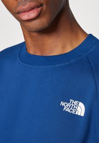 Sudadera de algodón azul con cuello redondo y puños acanalados. Presenta un logo blanco en el lado izquierdo. Textura suave, diseño ajustado.