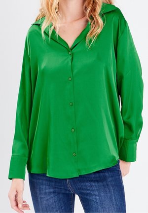 Femme portant un chemisier vert vif à boutons avec manches longues et un jean bleu, vue du cou jusqu'aux hanches sur un fond blanc.