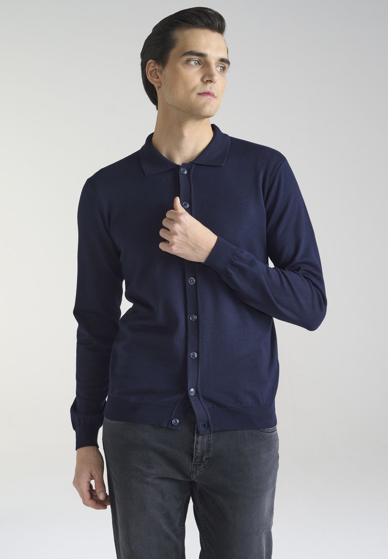 Granatowy kardigan wykonany z miękkiego materiału dzianinowego, z kołnierzykiem i zapięciem na guziki, z pasującymi guzikami. Założony z ciemnoszarymi spodniami.