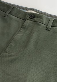 Vihreät ruutukuvioiset chinot nappisuljennalla, joissa on sileä rakenne, tasainen etuosa ja vyölenkit. Varren sisäpuolella on kontrastiväri vaaleansininen trimmi.