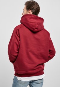 Felpa con cappuccio rosso realizzata in materiale morbido, caratterizzata da un cappuccio ampio e polsini a costine, abbinata a jeans azzurri chiari.