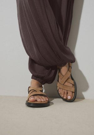 Nohy v semišových sandáloch svetlo hnedej farby s krížovými remienkami a čiernymi podrážkami, v kombinácii s voľnými tmavohnedými nohavicami stiahnutými pri členkoch.