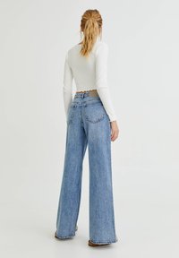 Jeans de mezclilla azul de pierna ancha con una cintura alta, que presentan un acabado limpio y sutiles detalles de costura. Combinados con una blusa blanca de manga larga.