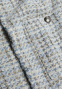 Tissu tweed tissé bleu clair et crème avec des accents noirs, featuring un bouton décoratif argenté et un détail de bord à franges.