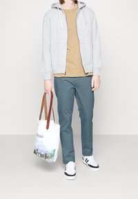 Grå zip-up hoodie, brun t-shirt, turkosa byxor, vita och svarta sneakers, samt en vit tote-väska med bruna remmar med en illustration.