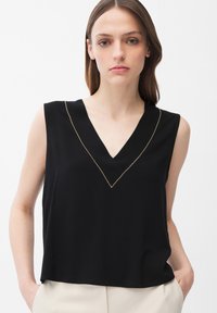 Joven con cabello castaño lacio que lleva una blusa negra sin mangas con escote en V y ribete de cuentas doradas, pantalones beige, manos en los bolsillos, fondo neutro.