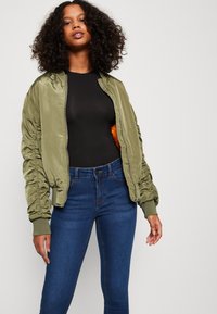 Casaco bomber de satin verde-oliva com mangas franzidas, combinado com uma blusa preta e jeans skinny azul-escuro. Apresenta um fecho com zíper e punhos canelados.