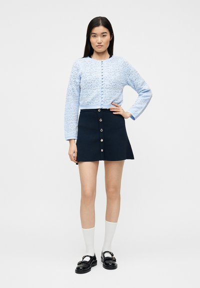 Γυναίκα που στέκεται, φορώντας γαλάζια δαντελένια ζακέτα με κουμπιά, σκούρο μπλε κουμπωτή φούστα, λευκές κάλτσες και μαύρα παπούτσια Mary Jane σε λευκό φόντο.