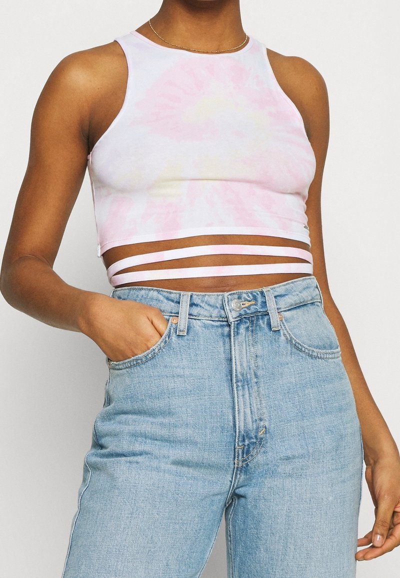 Top corto de tirantes blanco con efecto tie-dye rosa, combinado con unos vaqueros de cintura alta en azul claro. Tejido suave, diseño ajustado y estilo minimalista.