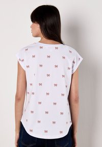 Una joven de espaldas, que lleva una camisa blanca con pequeños patrones de lazo rosa. Está de pie frente a una pared sencilla, mostrando un estilo casual.