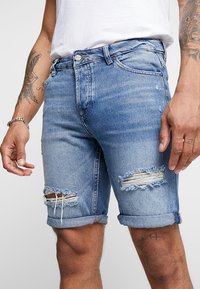 Shorts en jean de couleur bleu clair avec une finition délavée. Présentent des bords effilochés et des déchirures au niveau des genoux. Design classique à cinq poches, ourlets repliés.