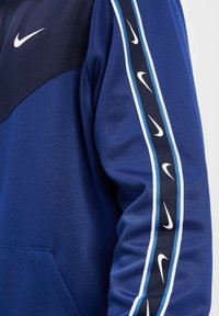 Zila un tumši zila kapuce ar pusi-zipa, kurai gar piedurknēm ir baltas Nike emblēmas, un priekšā ir kangaru kabata. Gluda auduma tekstūra.