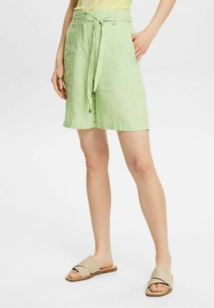 Femme portant un short taille haute vert clair avec une ceinture à nouer et des sandales beiges à enfiler, debout avec une main dans la poche.